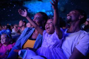A little girl smiling and holding her arms up while sat with her mum and dad in a theatre