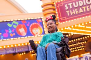 Guest in a wheelchair wearing Minnie Mouse ears smiling beneath the Studio Theater marquee at Production Courtyard, Disney Adventure World