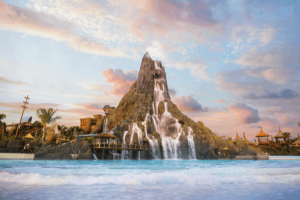 A wide shot of a massive artificial volcano with cascading waterfalls flowing into a large, wavy blue pool, under a pastel pink and blue sunset sky with scattered clouds