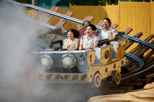 Riders smiling and cheering aboard a rustic mine cart roller coaster emerging from a cloud of mist