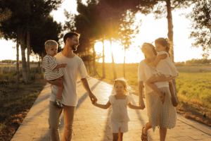 A happy family of five holding hands and carrying young children while walking along a paved path at sunset