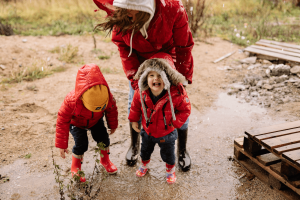 Two young children in red raincoats laugh and play in a muddy puddle while an adult in a red coat bends down beside them, illustrating a rainy day out with the family