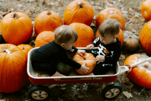 Two toddlers in skeleton costumes sit in a red Radio Flyer wagon at a pumpkin patch, holding a pumpkin between them.