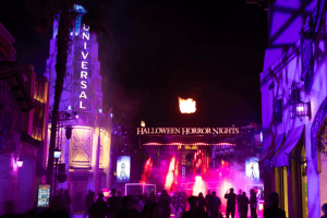 A crowd of guests walking toward the brightly lit Halloween Horror Nights entrance gate with bursts of fire in the night sky