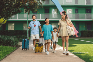 A family smiling and walking with their rolling luggage outside a green resort building