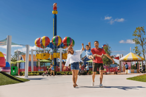 A mother and father swinging their young son by his arms while walking in front of a Peppa Pig balloon ride at a theme park