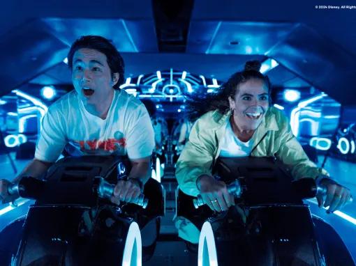Two young adults sat on motorcycles shaped vehicles TRON Lightcycle. They are leaning forward gasping with shock. 