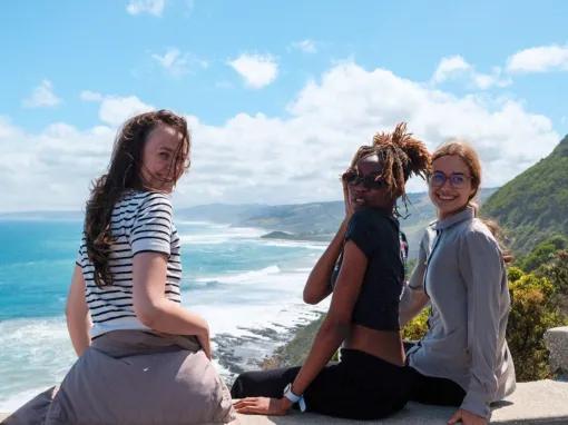 Three people smiling, sitting on the edge overlooking the ocean at a lookout