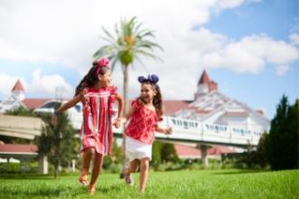 Two little girls wearing Mickey Ears running over grass while holding hands and laughing, with Disney's Grand Floridian in the background