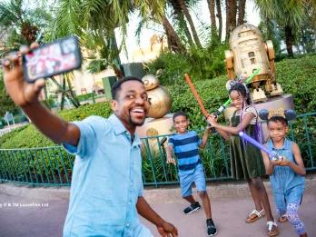 A father and three children taking a selfie posing with lightsabers in front of the Star Wars statues