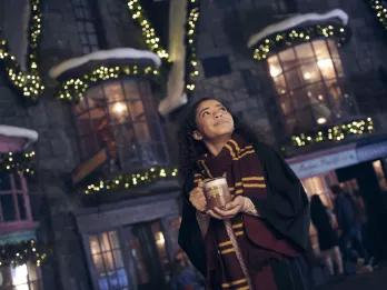 Christmas in Orlando A young woman wearing a scarf and wizard robes drinking Butterbeer in front of decorated buildings
