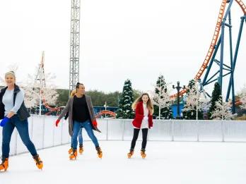 4 adults ice skating outdoors in front of a rollercoaster