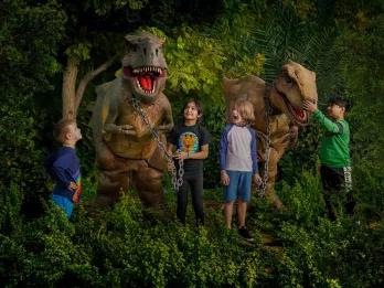 4 children standing in some foliage with two dinosaur animatronics