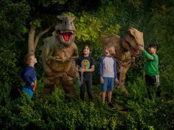 Dino Mania at Dubai Parks and Resorts 4 children standing in some foliage with two dinosaur animatronics