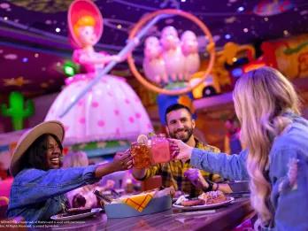 3 people sat at a table clinking mason jars full of drinks. They are sat in front of a large figure of Bo Peep from Toy Story
