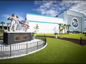 Apollo V Center at Kennedy Space Center A statue of three astronauts holding an American flag in front of a building with a large sign reading Apollo NASA
