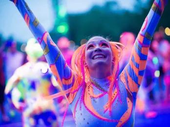 A female performer in a neon blue and orange leotard looking up to the sky with her arms raised