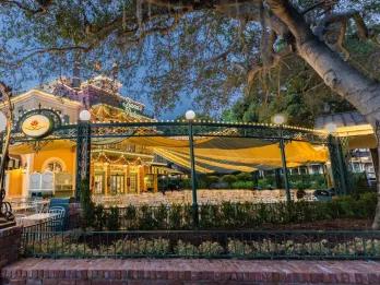 The exterior of a restaurant with white chairs and tables covered by a green and yellow striped awning and green ironwork