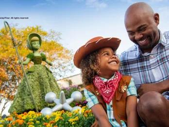 EPCOT International Flower & Garden Festival A young boy dressed as a cowboy smiling up at his dad. They are both sat in front of a topiary shaped like Bo Peep from Toy Story