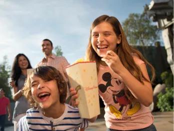 Girl wearing pink Mickey Mouse top holding popcorn bucket in one hand and a piece of popcorn in the other, laughing with a young boy who is next to her. In the background is a couple smiling and a large star wars robot.