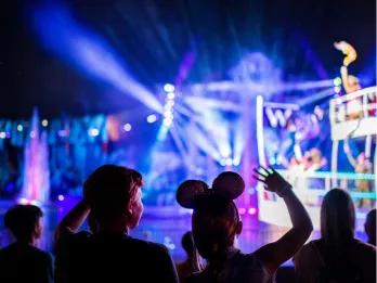 Behind two children watching Fantasmic! One girl wearing mickey ears and one boy with one of each of their hands in the air. In the blurry background there are multicoloured lasers shining, purple and blue lighting, fountains and the front of a white ferry boat holding Disney characters.