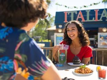 A woman eating a cupcake and drinking a cocktail from a pouch sitting across a table from someone