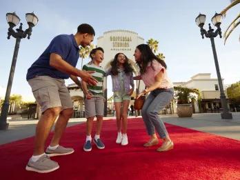 Family jumping on the red carpet in front of the large white stone entrance sign with Universal Studios Hollywood logo on. The family has dad on the right, one girl and one boy in the middle jumping and the mum crouched on the right.