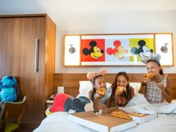 Three girls sat on a hotel bed eating slices of pizza from a pizza box