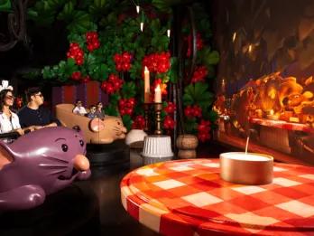 Guests sat in a rat shaped vehicle with 3D glasses on. Next to them on the right is a giant bush with large flowers, a giant table with a red and white table cloth, giant candles and a screen with the ratatouille characters on. 