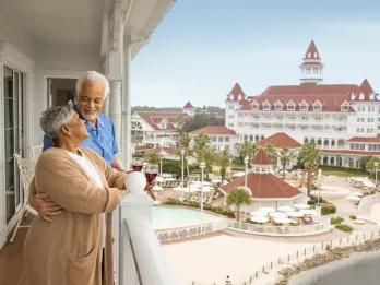 An elderly couple standing on a balcony looking out on a white and red Victorian-style building