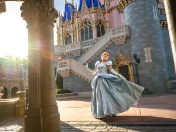 Cinderella by Cinderella's Castle  Princess Cinderella has her blonde hair in a bun and her blue ballgown on and is dancing underneath a stone archway in front of the pink and blue castle.