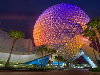 Epcot Spaceship Earth Giant white ball with purple and orange lighting. Beneath there are palm trees.