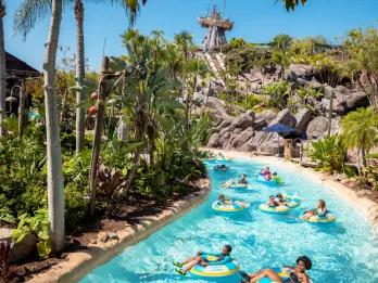 Typhoon Lagoon Lazy river, long pool with guests relaxing/lounging on rubber rings in the clear water. In the background there are rocks and trees, and a statue of a shipwreck on top of a tall rock.