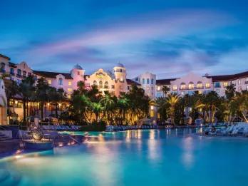 White large hotel in the background with palm trees in front and a huge swimming pool.