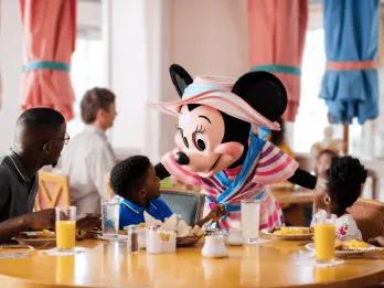 Minnie Mouse greeting two young children and their dad while they are sat round a table eating breakfast