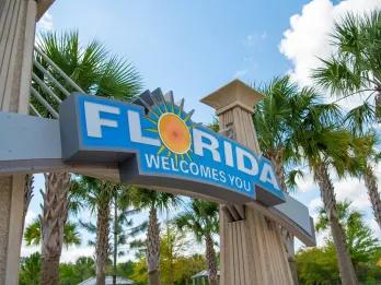 A blue sign with white text which says 'Florida Welcomes You' with palm trees in the background.