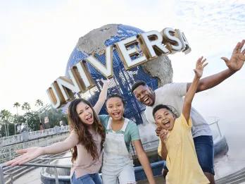 A family of four stood in front of a big circular statue of the globe with white capital letters on spelling UNIVERSAL. The family are smiling and have their arms spread wide. There is a couple with one daughter and one son.