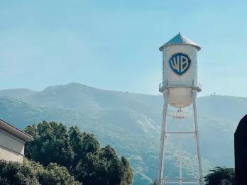 Warner Bros. Studio White water tower with blue shield shaped logo with letters WB on. In the background there is a large tree and big mountains with a blue sky overhead.
