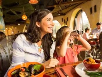 A woman and child dining in the Animal Kingdom Lodge, the mother is smiling looking off camera through a window and the little girl is also smiling looking in the same direction with binoculars held against her eyes. The mother has a bowl of seafood in front of her whereas the girl has a plate with chicken curry, rice and green beans.