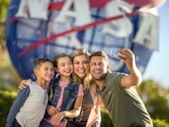 Family of four, a man holding up a phone to take a selfie with wife and two kids, one boy and one girl. They are stood in front of a large sign, shaped like the earth, circular with red stripes over and the letters in white spelling NASA.