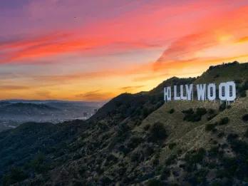 Large mountains surrounding a city. There is a large sign with capital letters spelling out "HOLLYWOOD". In the background there is a sunset with pink and orange colouring.