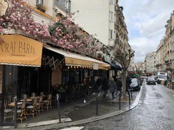 Paris restaurant A wet road in Paris lined with parked cars and the outside of s restaurant which is named "Vrai Paris". Above the beige cover there are pink flowers and below there are tables with chairs.