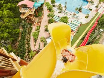 Birds eye view of man dropping down the top of a yellow vertical drop slide, beside there is a red which is smaller. There are pools below and walkways with people queueing for smaller waterslides.