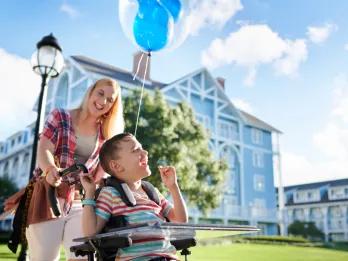 Young boy in a wheelchair that has a blue mickey shaped balloon tied to it. Behind the mum is pushing him and they are both smiling. Behind there are tall buildings which are pastel blue with white decorative beams.