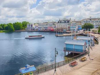 Boardwalk with a large lake to the left. There are stalls with stripey covers and a small boat heading towards the dock. There are buildings lining the boardwalk of different pastel colours.