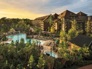 Large brown building in the middle of a forest type landscape. There are also two pools, one large and surrounded by rows of sunbeds and the other smaller. 