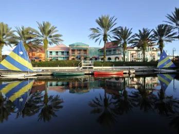 A lake with small rowboats and sailing boats in it. Behind there are multiple buildings of various pastel colours.