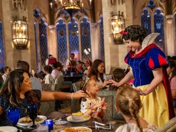 Mum and two girls sat a table eating. The mum is touching one of the girls hair as the little girl beams up at Princess Snow White who is at their table. Snow White is looking down smiling with a shocked expression. The small girl looks so surprised and happy. In the background there are tall windows on the brick walls and a large chandelier.