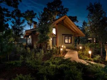 Modern cabin with light wood design, surrounded by trees, which has a small stone path leading up to it and short lamps leading the way.