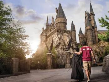 Hogwarts Castle A man wearing a red Griffindor shirt holding hands with a child in a black cloak and a wand. They are walking along an empty walkway in front of Hogwarts Castle.